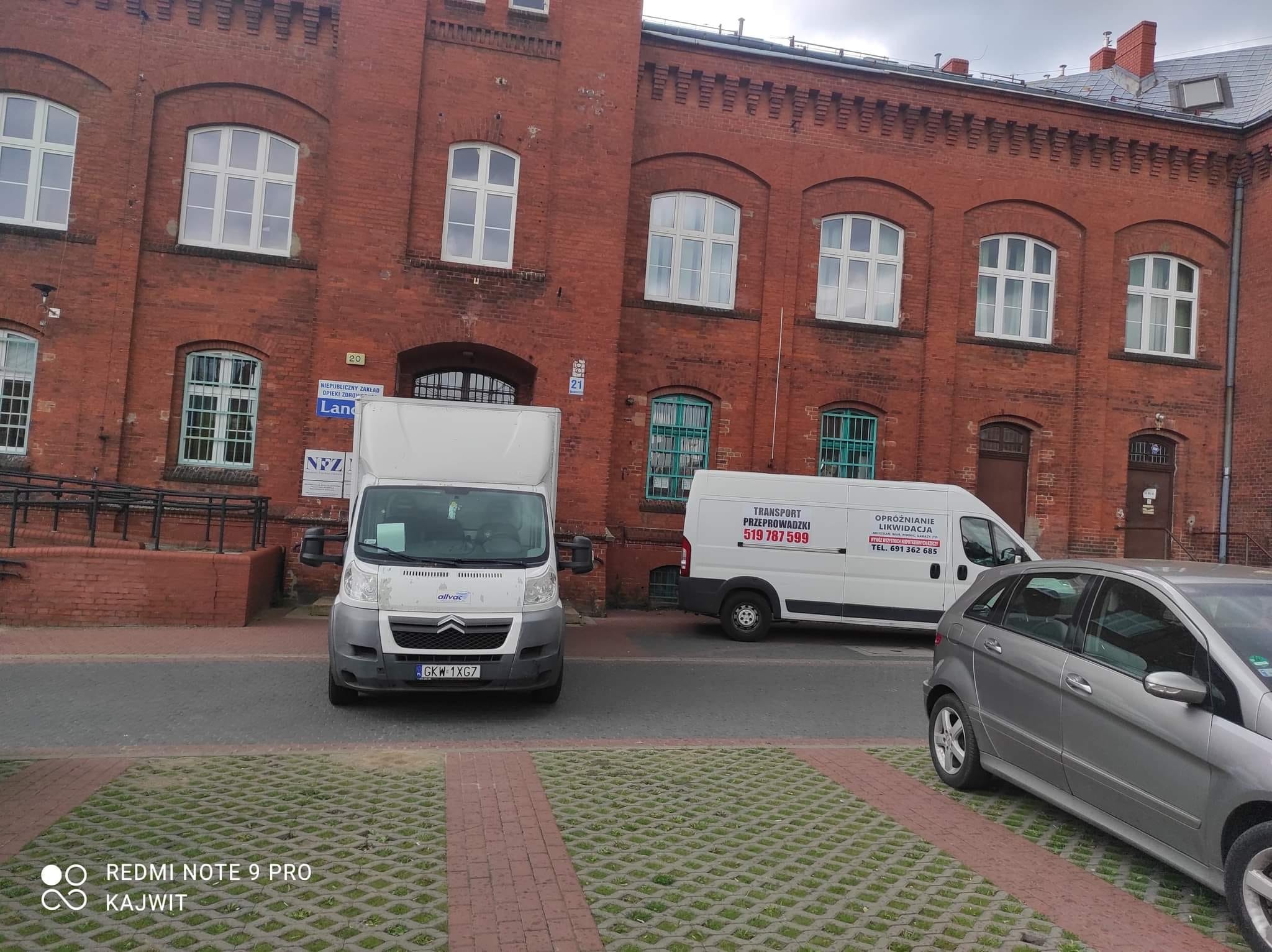 Dwa białe busy zaparkowane przed budynkiem z czerwonej cegły; jeden z napisami 'TRANSPORT PRZEPROWADZKI' i numerem telefonu, drugi z logo firmy.