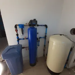 Zestaw do uzdatniania wody: niebieski filtr narurowy, pionowy niebieski zbiornik, beżowy zbiornik hydroforowy, instalacja z niebieskich rur w surowym wnętrzu.