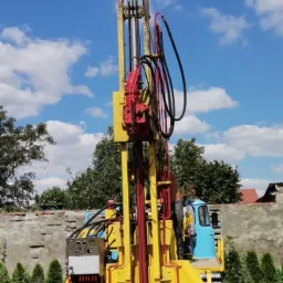 Żółto-czerwona wiertnica na podwoziu ciężarówki, z operatorem w kabinie, na tle muru z jasnego kamienia i błękitnego nieba z obłokami.