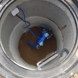 Wnętrze betonowego szamba z zainstalowaną pompą, niebieskimi przewodami i elementami instalacji, widoczne drabinki ze stali, biała puszka elektryczna na ścianie szamba.