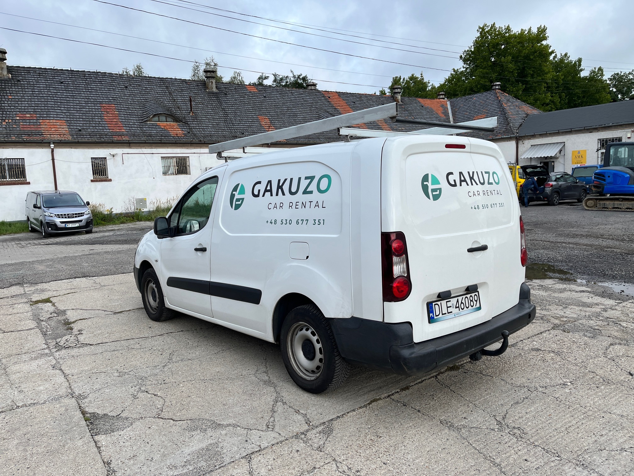 Biała furgonetka z logo Gakuzo Car Rental, zaparkowana na betonowym placu w Legnicy, z widocznym bagażnikiem dachowym i numerem rejestracyjnym DLE 46089.