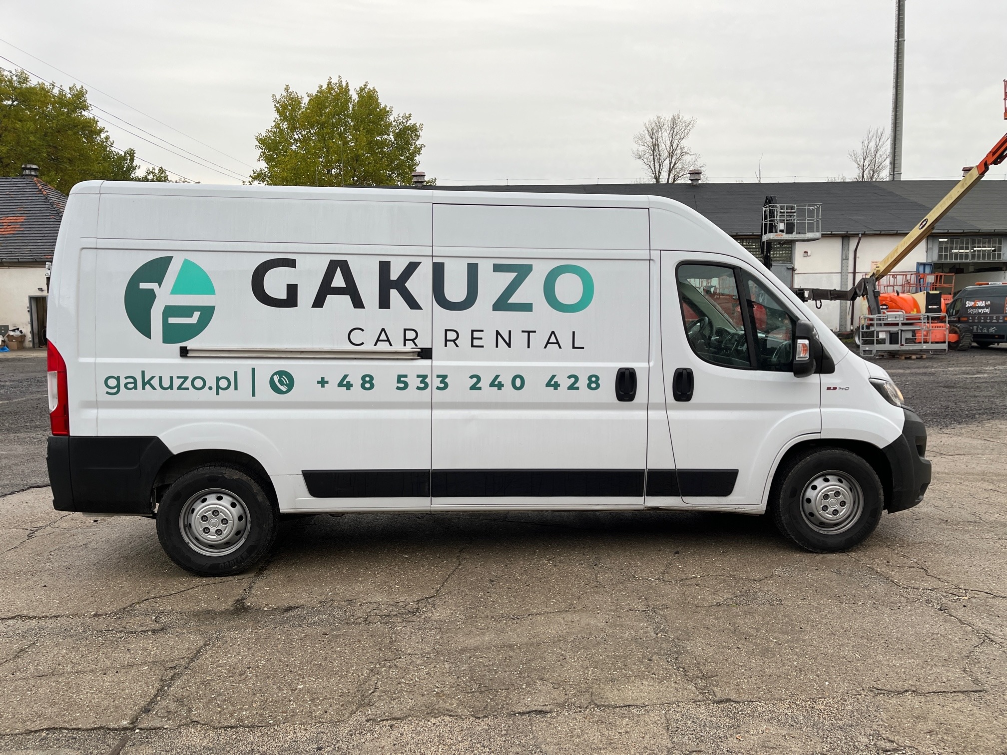 Biały samochód dostawczy z logo Gakuzo Car Rental i numerem telefonu zaparkowany na betonowym placu, w tle budynek i pomarańczowy podnośnik koszowy.