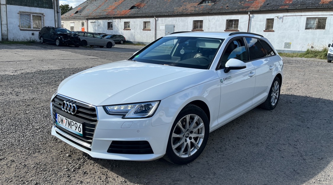 Białe Audi A4 Avant na żwirowym parkingu z widocznym logo Cakuzo Car Rental na przednim błotniku, z uszkodzonymi samochodami w tle i budynkiem z czerwonym dachem.