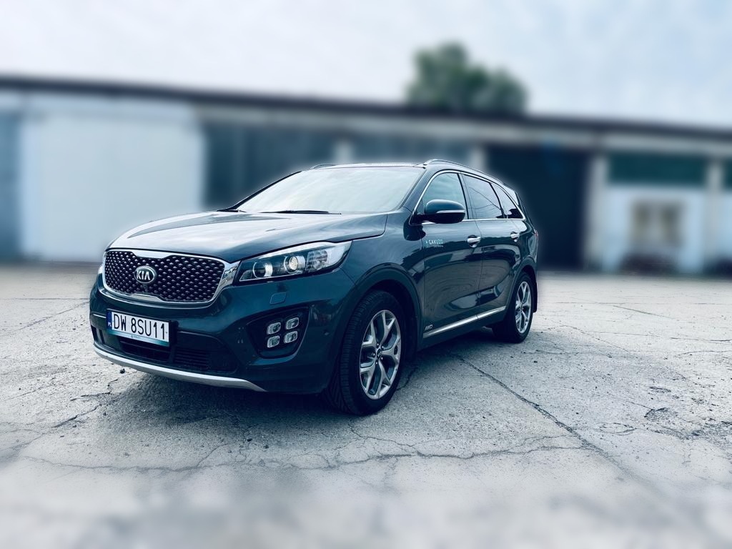 Ciemnozielony SUV Kia Sorento z widoczną tablicą rejestracyjną DW 8SU11, zaparkowany na betonowym placu przed budynkiem magazynowym z rozmytym tłem.