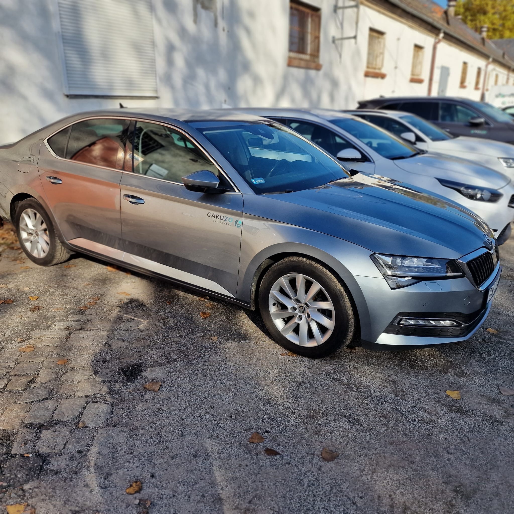 Szary samochód osobowy marki Skoda Superb z logo wypożyczalni Gakuzo Car Rental zaparkowany na placu z kostki brukowej, w tle biały budynek z roletami.