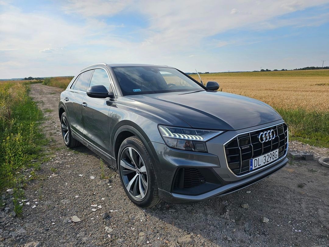 Szary SUV Audi Q8 na polnej drodze, z widocznym polem zboża w tle i numerem rejestracyjnym DL 3298G.