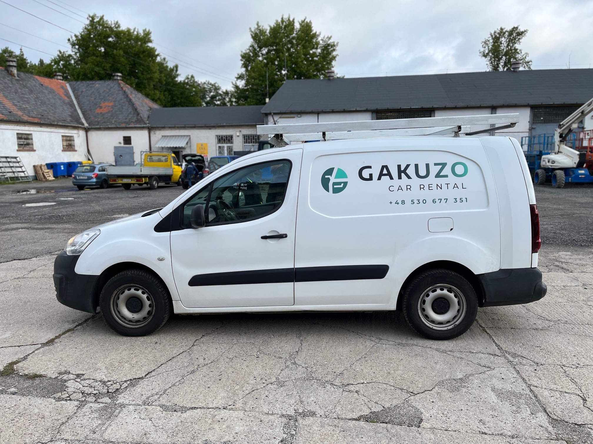 Biały samochód dostawczy z logo Gakuzo Car Rental zaparkowany na betonowym placu, drabina na dachu, w tle budynki gospodarcze i inne pojazdy.