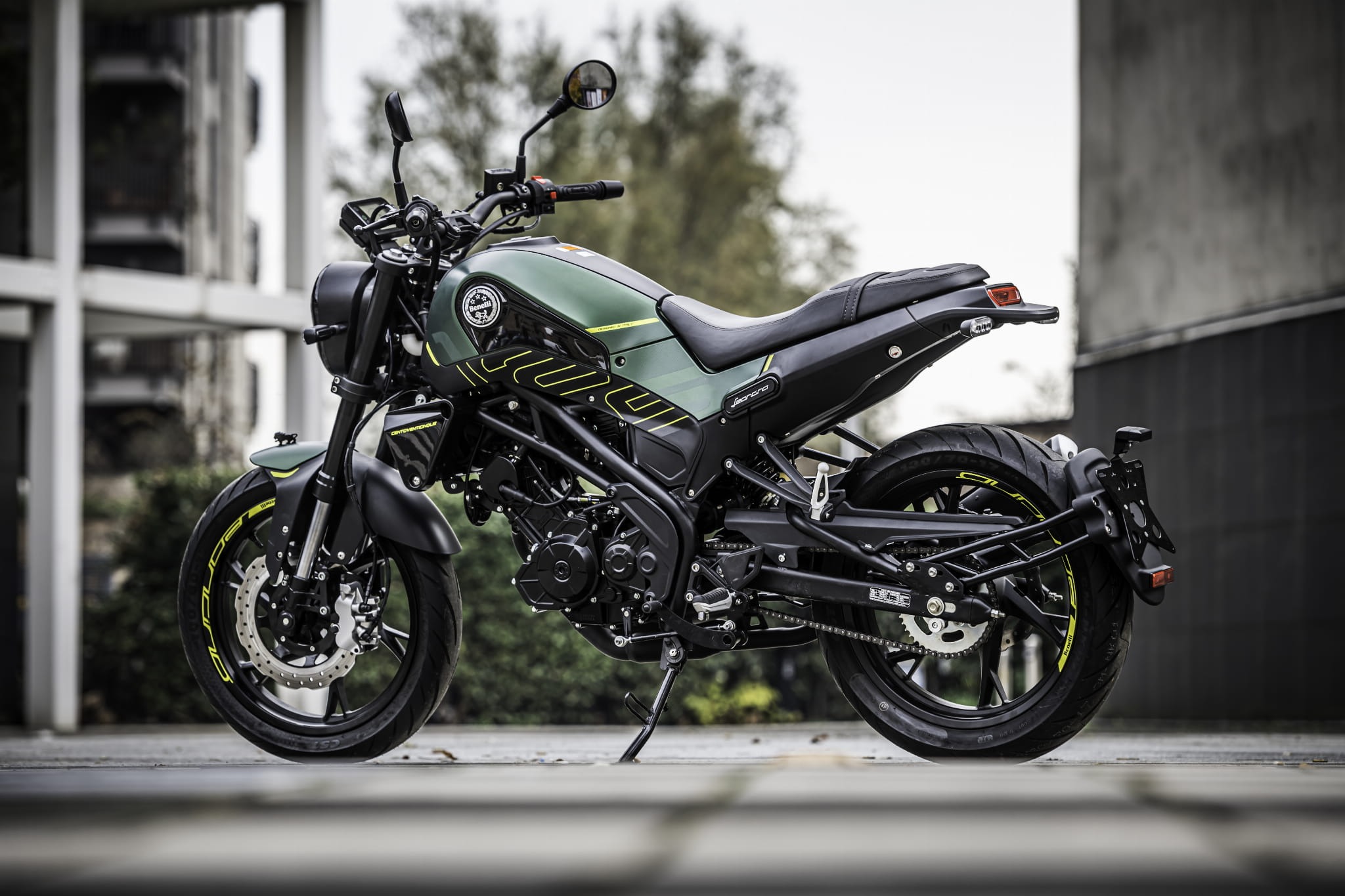 Nowoczesny motocykl w kolorze oliwkowej zieleni z czarnymi akcentami i neonowymi żółtymi detalami, zaparkowany na tle budynku o architekturze modernistycznej.