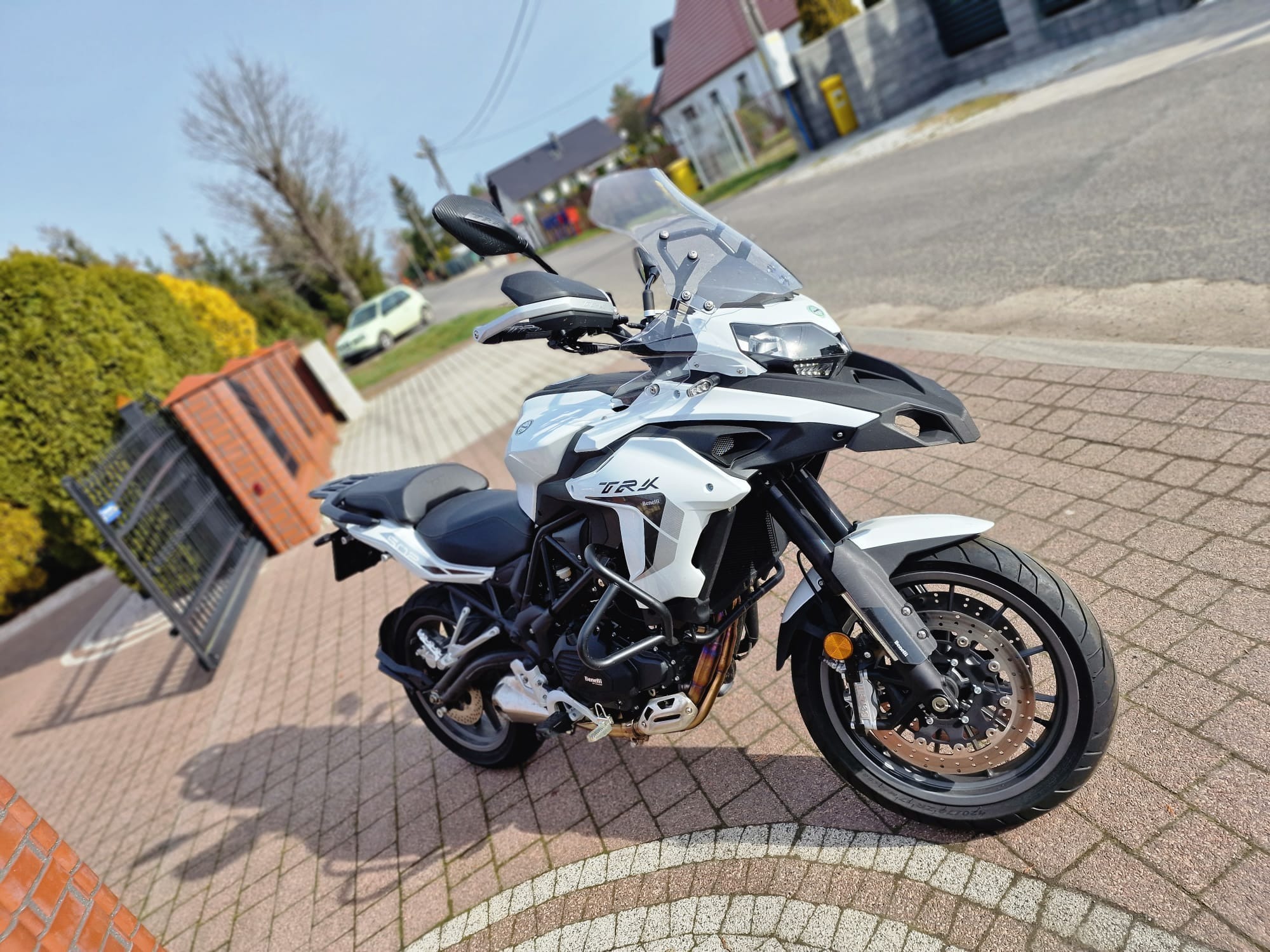 Biały motocykl turystyczny Benelli TRK 502 X zaparkowany na kostce brukowej przed domem w słoneczny dzień.