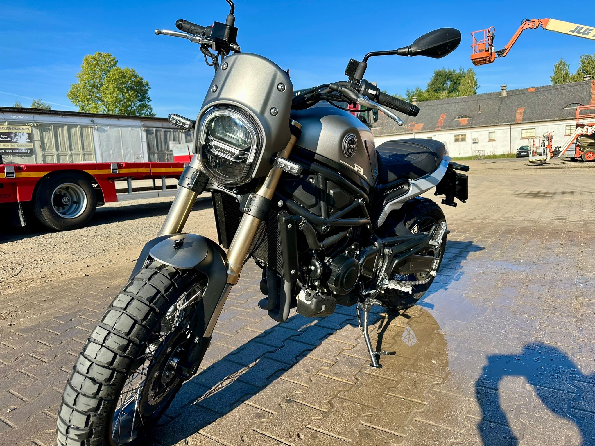 Szary motocykl Benelli Leoncino Trail z oponami terenowymi, zaparkowany na kostce brukowej, w tle budynek i podnośnik koszowy.