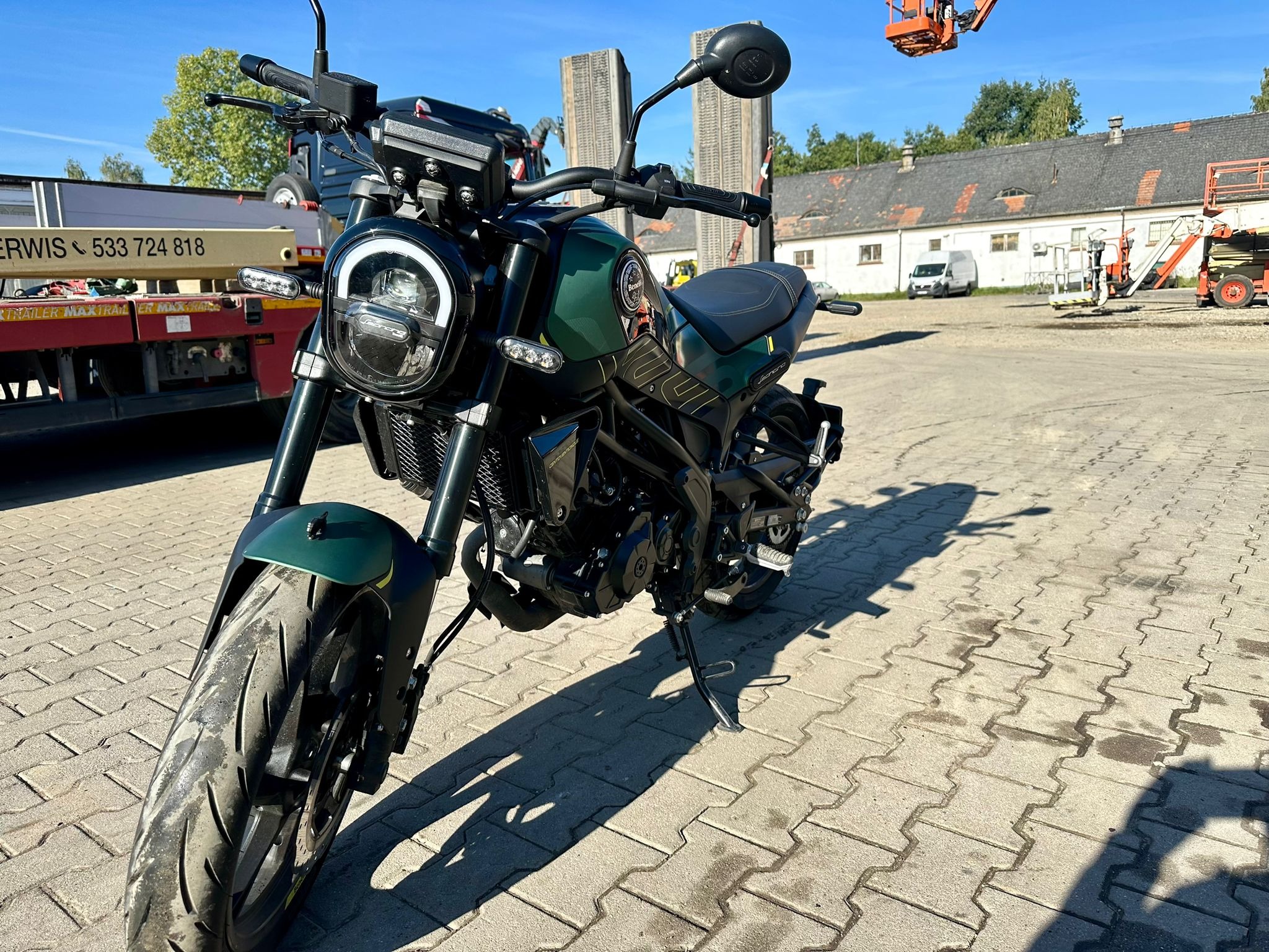 Motocykl typu naked bike w kolorze ciemnej zieleni, zaparkowany na brukowanym placu, z widocznym pojazdem pomocy drogowej w tle i podnośnikiem koszowym w oddali.