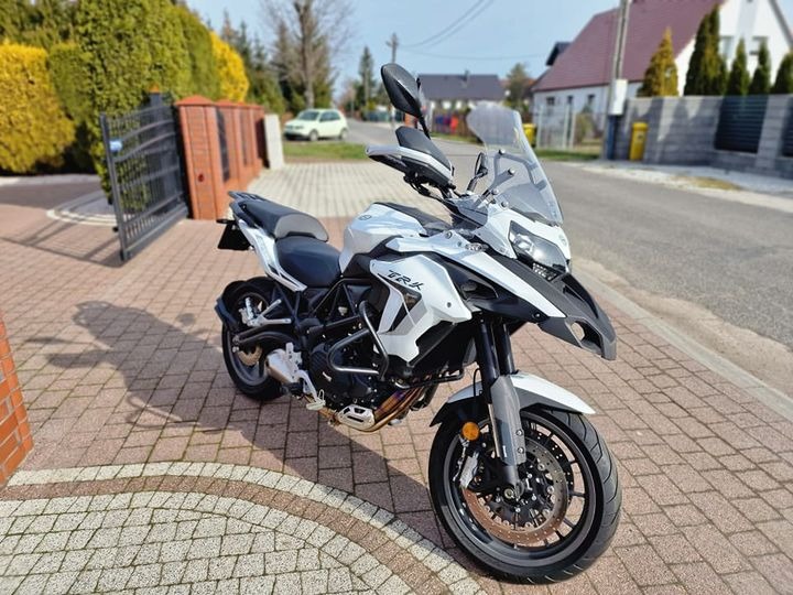 Biało-czarny motocykl turystyczny Benelli TRK na kostce brukowej przed domem z czerwonej cegły i zielonymi tujami.