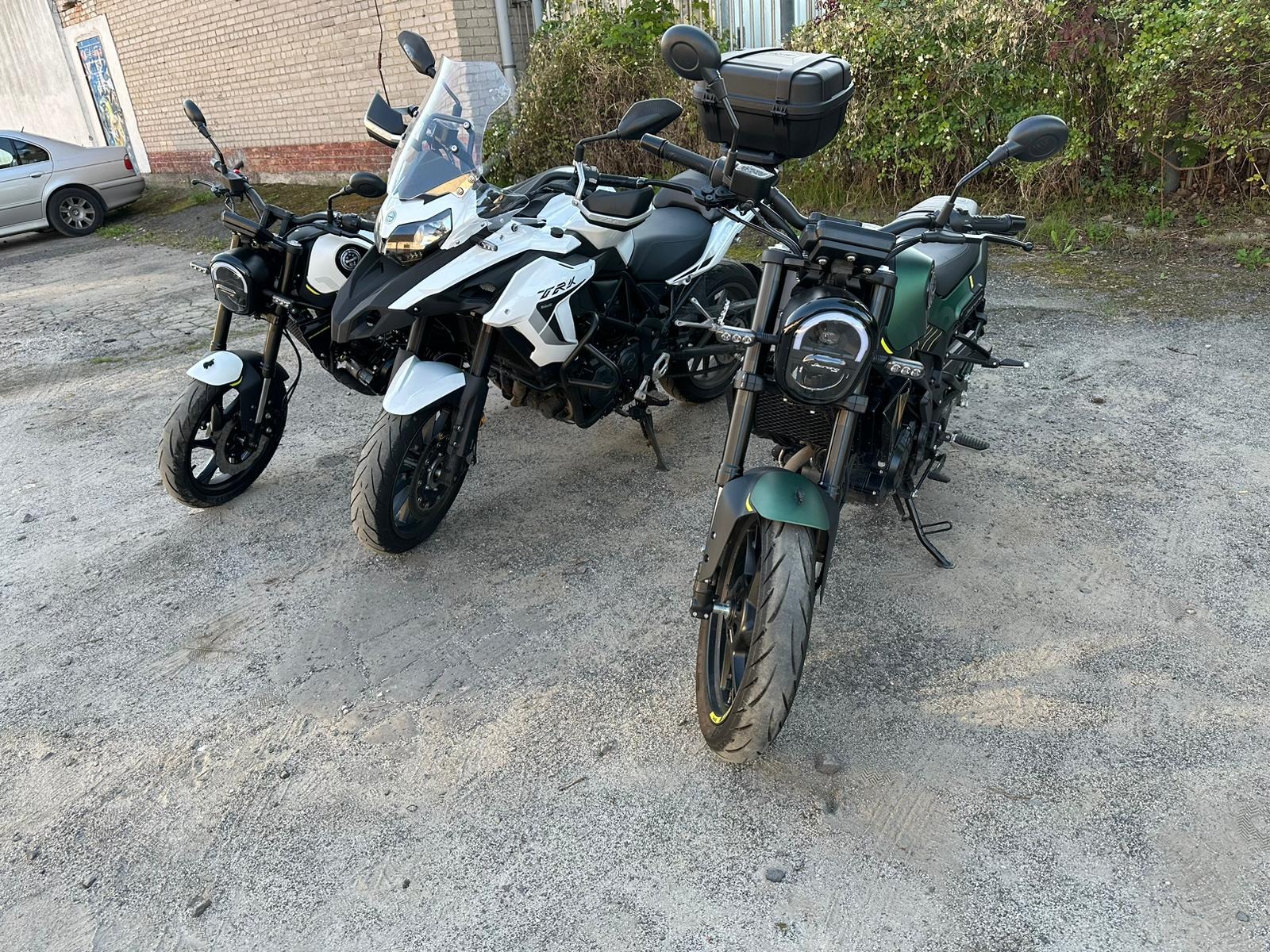 Trzy motocykle różnych modeli zaparkowane obok siebie na żwirowym podłożu, z zielonym motocyklem z przodu i białym motocyklem typu adventure w środku, a czarnym motocyklem typu scrambler z lewej...