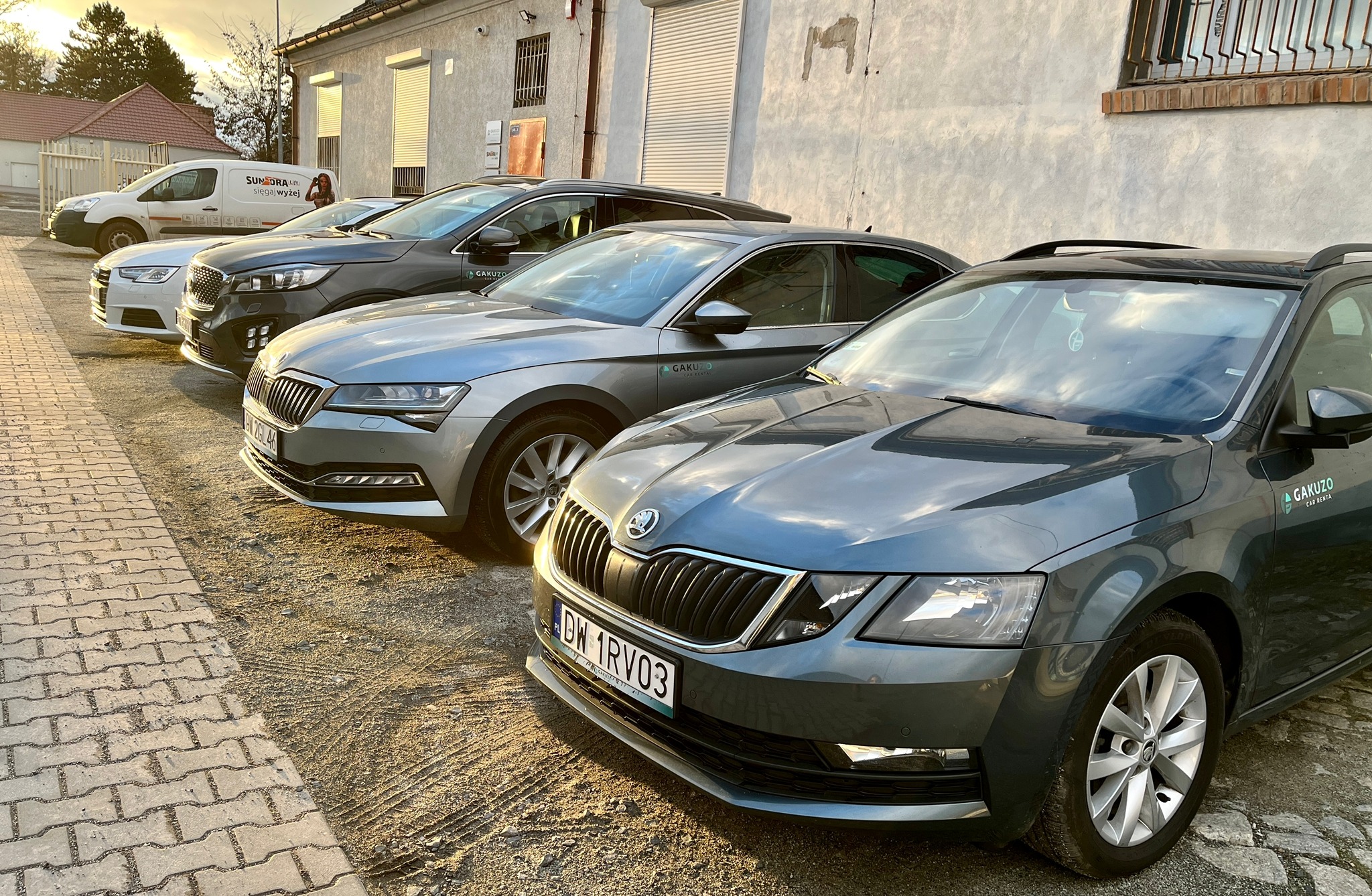Szereg zaparkowanych samochodów Skoda i innych marek, z logo wypożyczalni Gakuzo Car Rental na przednim samochodzie Octavia w kolorze szarym, na tle budynku z zamkniętymi roletami i wieczornym niebem.
