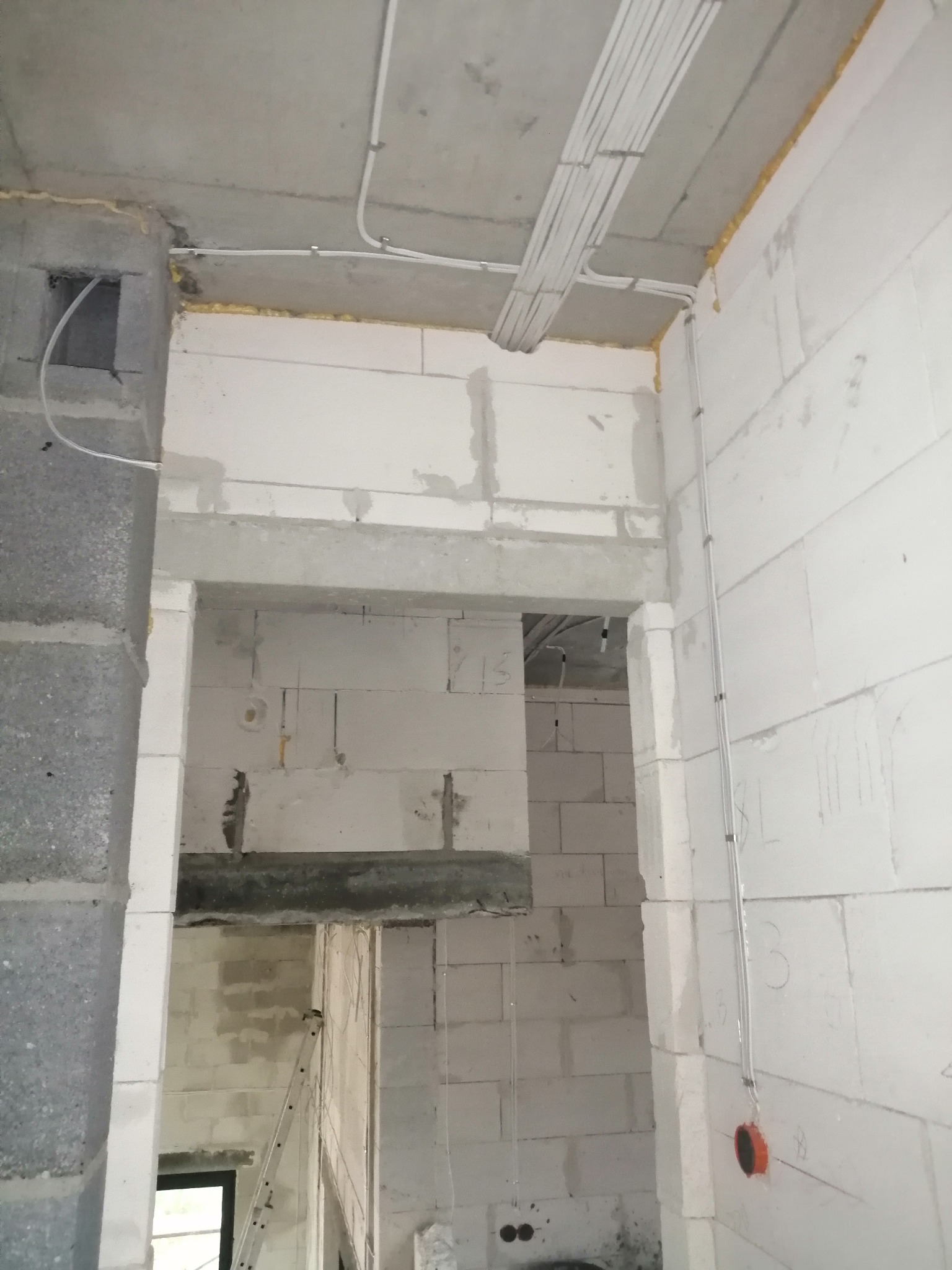 Wnętrze w trakcie budowy z widocznymi przewodami elektrycznymi przytwierdzonymi do sufitu i ścian, kable w peszlach, ściany z białych bloczków, widoczna drabina w tle.
