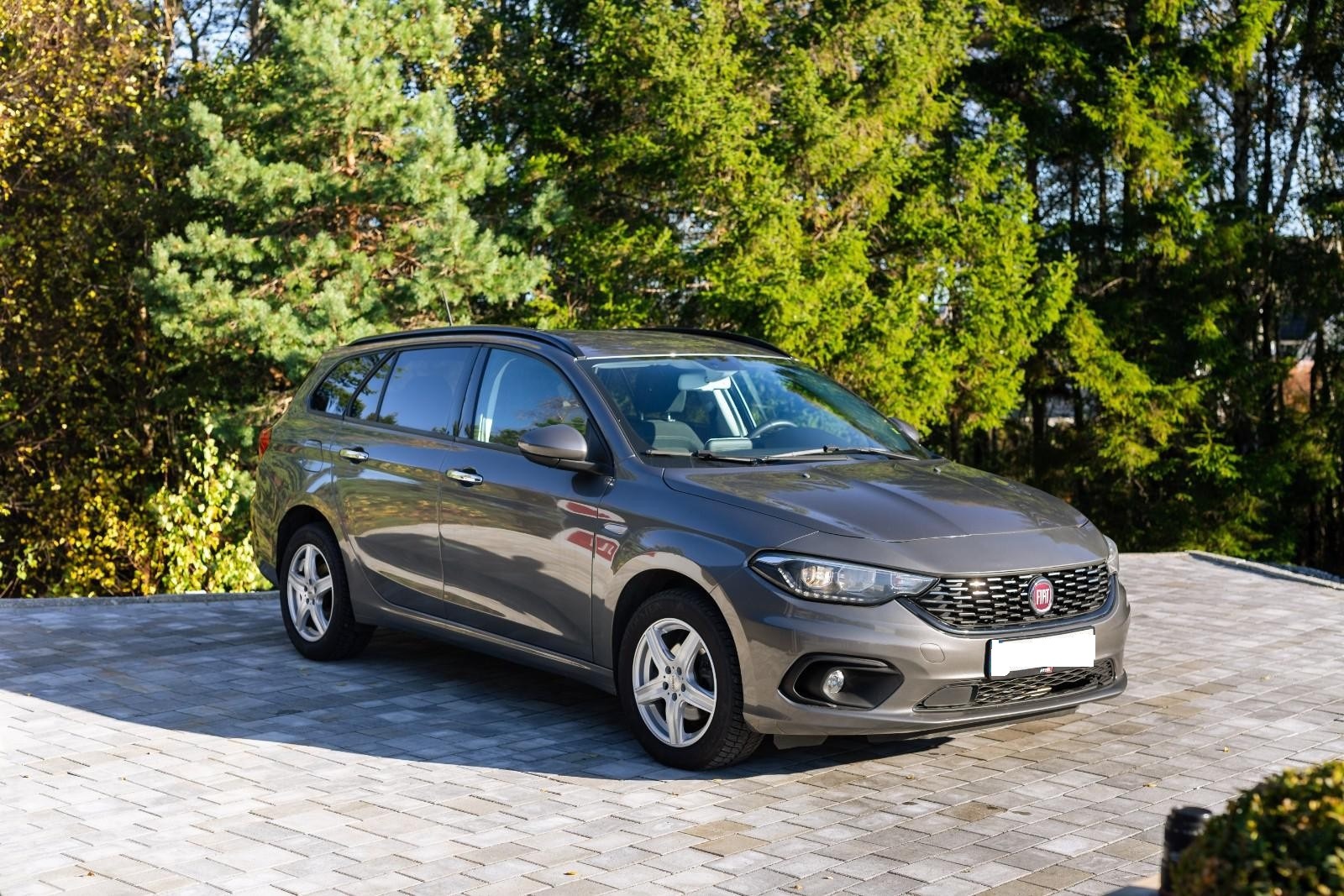 Szary Fiat Tipo kombi, zaparkowany na kostce brukowej z zielonym tłem drzew.