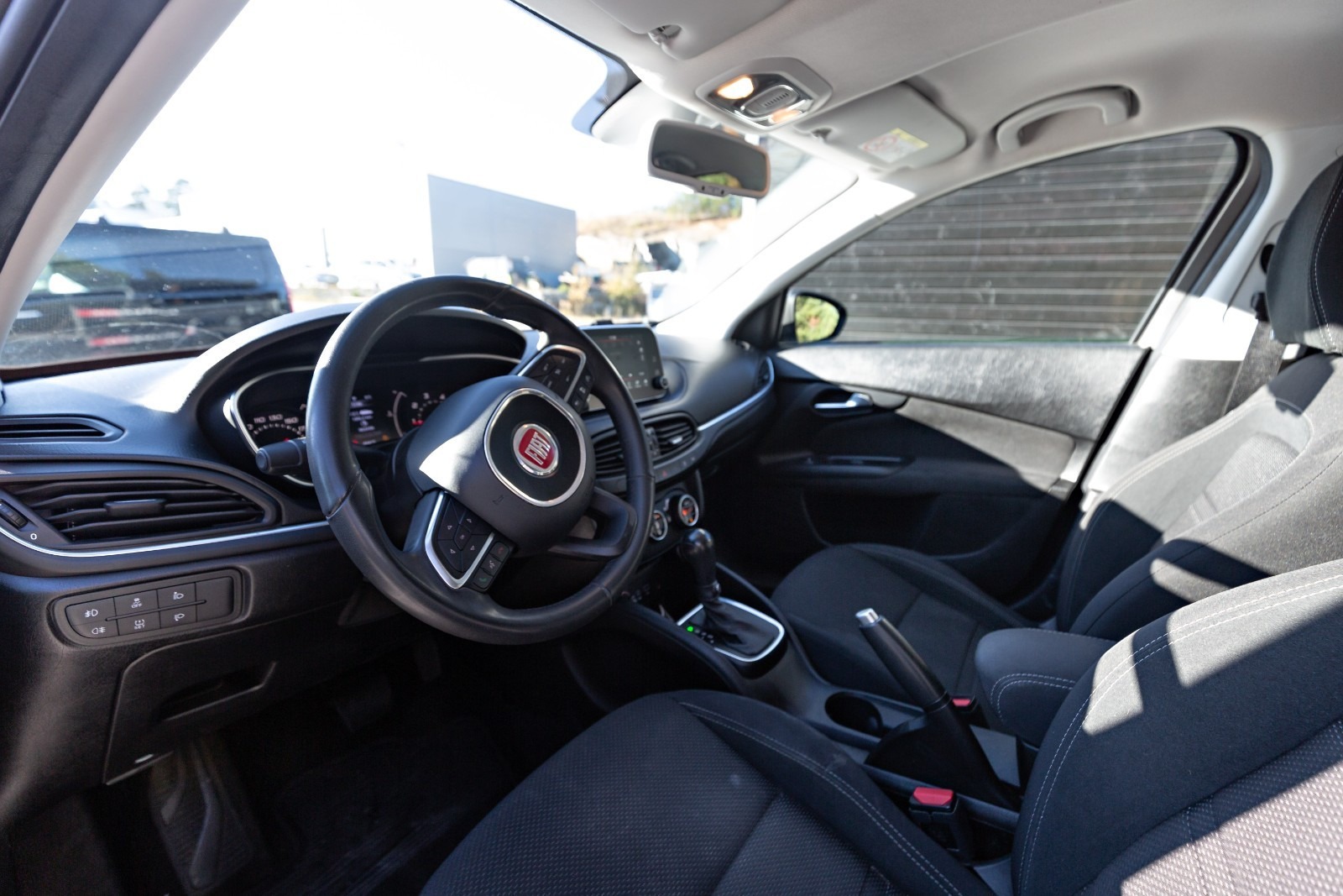 Wnętrze samochodu marki Fiat z widoczną deską rozdzielczą, kierownicą z logo firmy oraz fotelami z ciemną tapicerką.