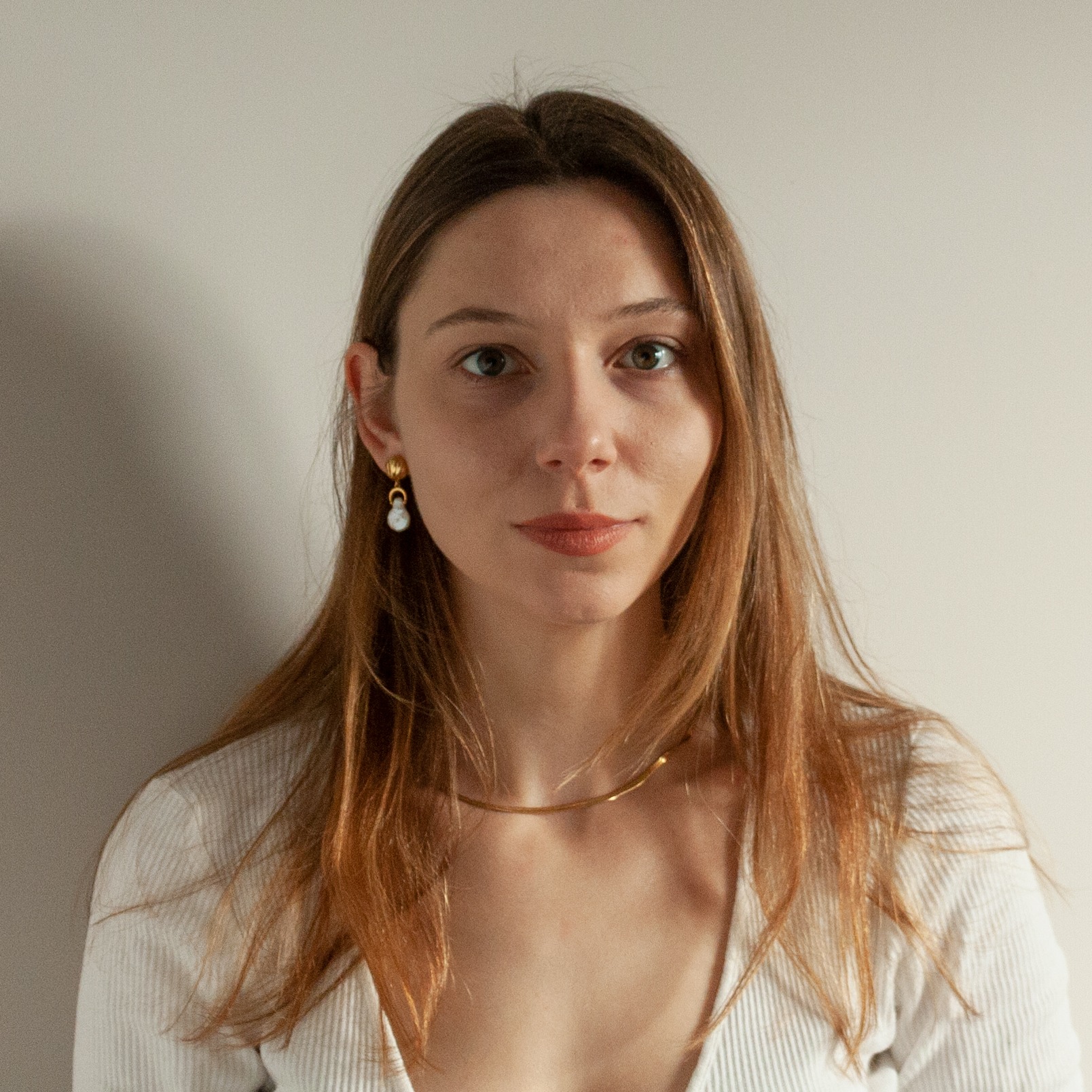 Portret kobiety z długimi, rudymi włosami, w białej bluzce i złotej biżuterii, sfotografowanej w Krakowie na jasnym tle z cieniem.