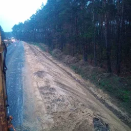 Żółty element koparki widoczny z perspektywy operatora na tle drogi w budowie, z ułożonymi stożkami ostrzegawczymi i lasem w tle, w pochmurny dzień.