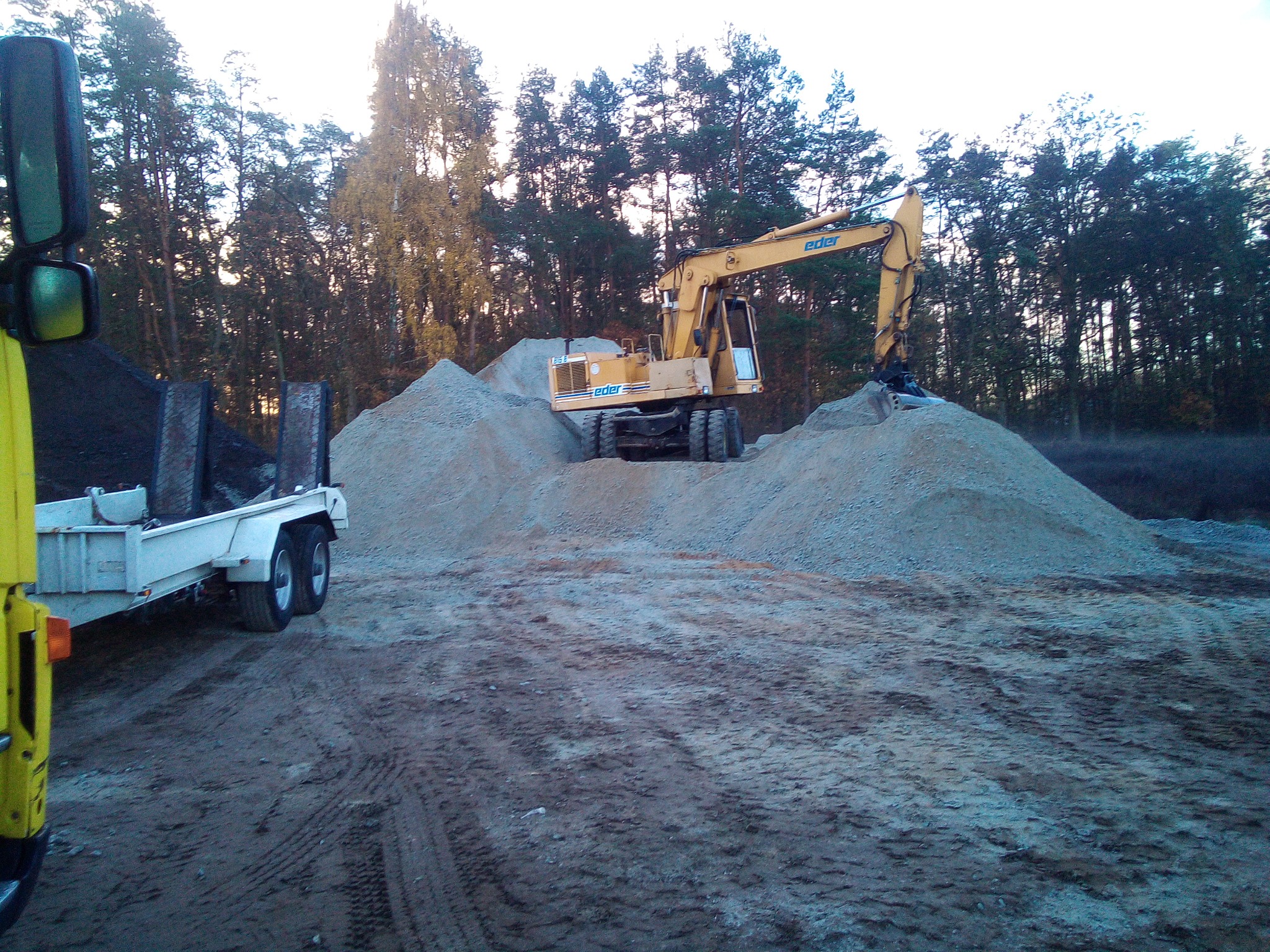 Żółta koparka Eder na nierównym terenie, obok przyczepa z czarnym ładunkiem, w tle las.