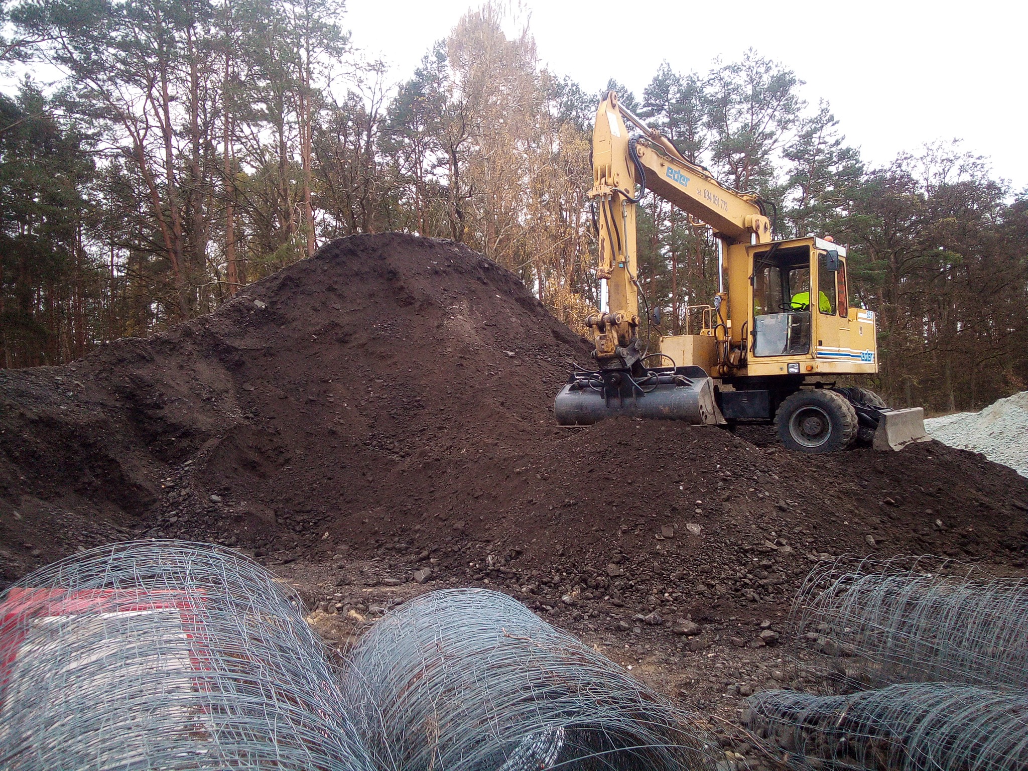 Żółta koparka Eder na tle sterty ziemi i lasu, z rolkami siatki ogrodzeniowej na pierwszym planie, w pochmurny dzień.