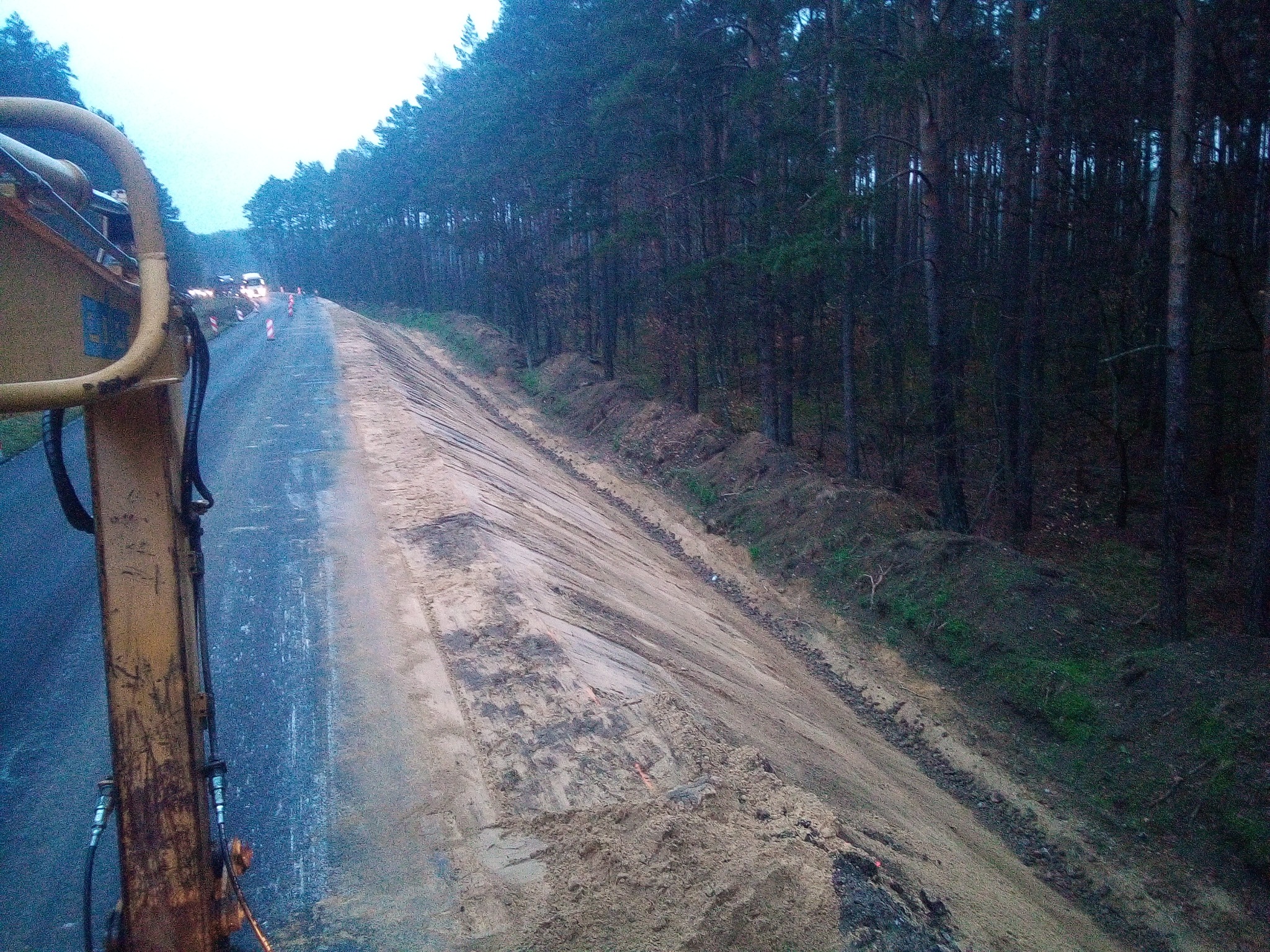 Żółty element koparki widoczny z perspektywy operatora na tle drogi w budowie, z ułożonymi stożkami ostrzegawczymi i lasem w tle, w pochmurny dzień.