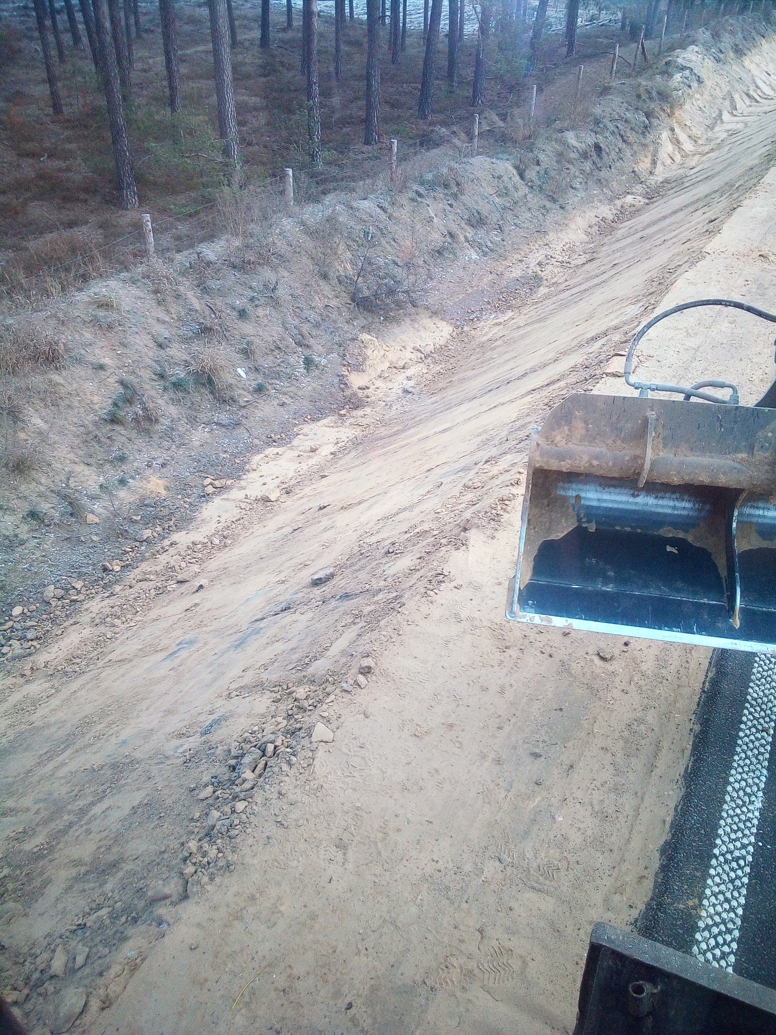 Widok z góry na wykopany rów w piaszczystym terenie, z widoczną krawędzią lasu w tle i fragmentem łyżki koparki w prawym dolnym rogu, obok widoczny fragment drogi z białym oznakowaniem.