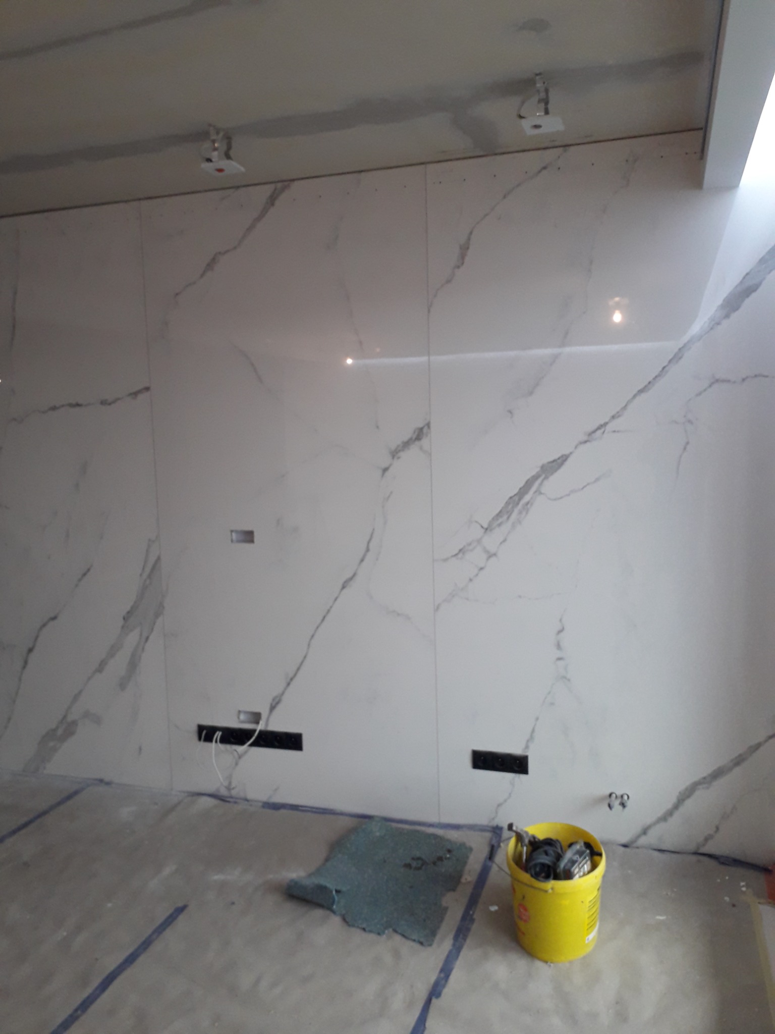 Ściana wyłożona białymi płytami imitującymi marmur, z widocznymi otworami na gniazdka elektryczne i puszkę, przy podłodze leży wiadro z narzędziami i kawałek materiału zabezpieczającego podłogę.