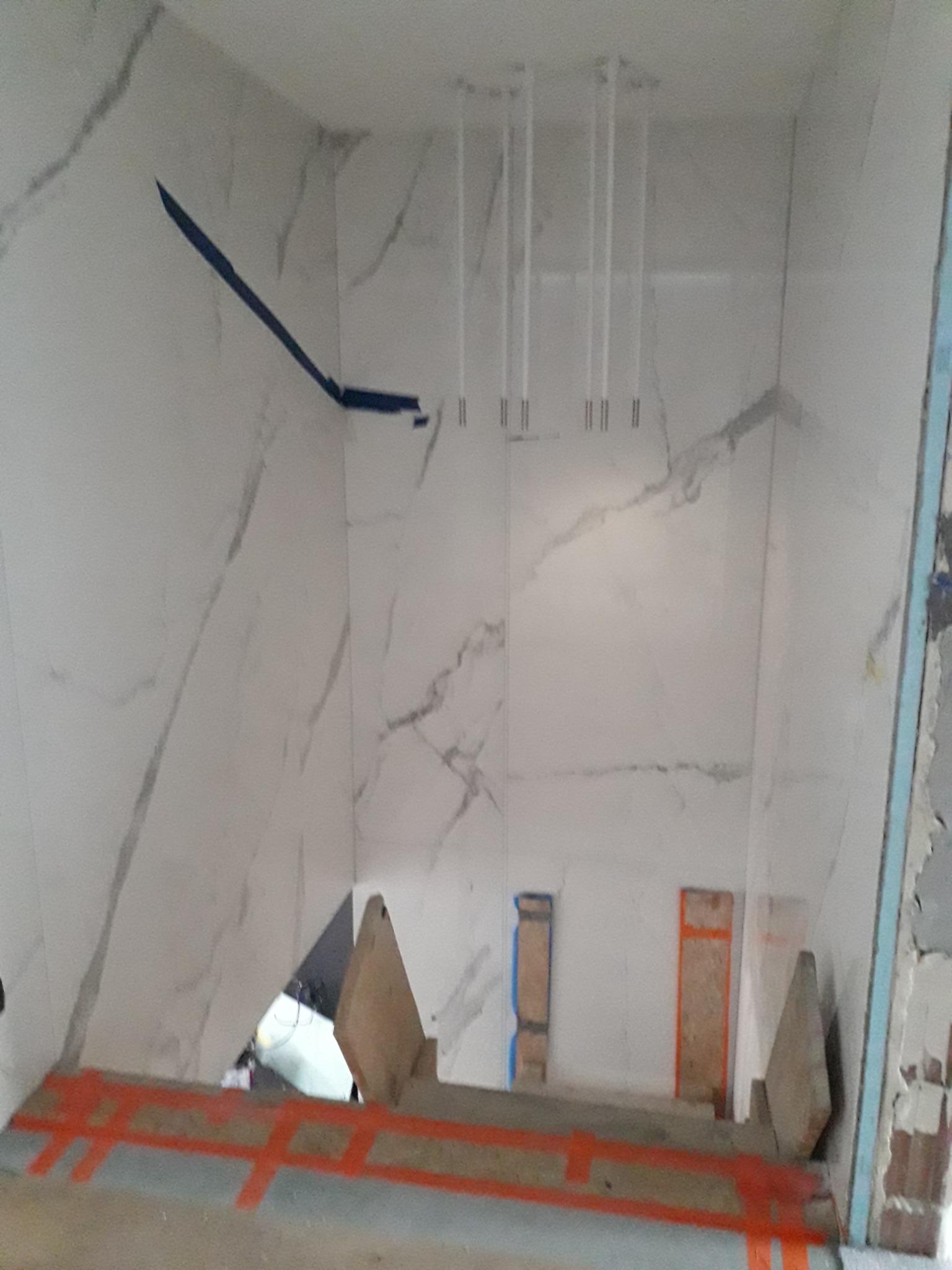 Wnętrze łazienki w trakcie remontu, widoczne białe płytki imitujące marmur na ścianach, zabezpieczone niebieską taśmą, przygotowane rury do instalacji oświetlenia sufitowego oraz zabezpieczona...