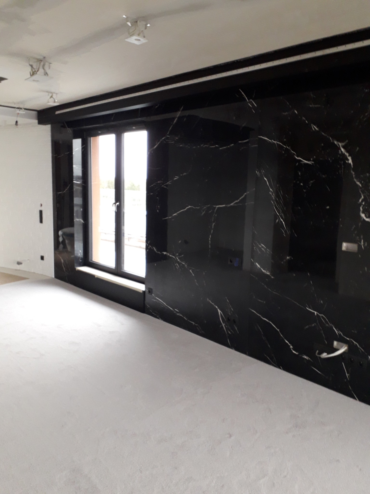 Ściana wykończona czarnym szkłem imitującym marmur z białymi żyłami, widok na okno, surowy stan podłogi i sufitu, widoczne instalacje elektryczne.