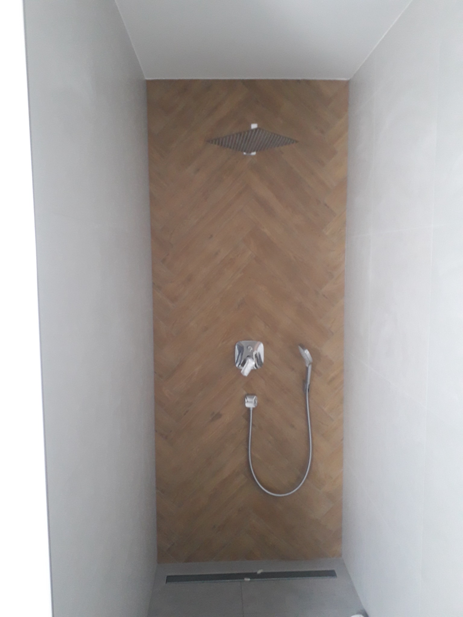 Kabina prysznicowa z deszczownicą i baterią podtynkową, wykończona płytkami imitującymi drewno ułożonymi w jodełkę, z odpływem liniowym w szarej posadzce.