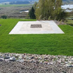 Widok z góry na świeżo założony trawnik z rolki, z centralnie umieszczonym kwadratowym paleniskiem wyłożonym kamieniami, otoczonym płytami chodnikowymi, na tle malowniczego krajobrazu z jeziorem...