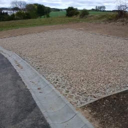 Utwardzony plac z kruszywa, ograniczony betonową obrzeżem i ażurowymi płytami, przylegający do asfaltowej drogi z betonowym korytkiem odwadniającym. W tle widoczne drzewa i pola.
