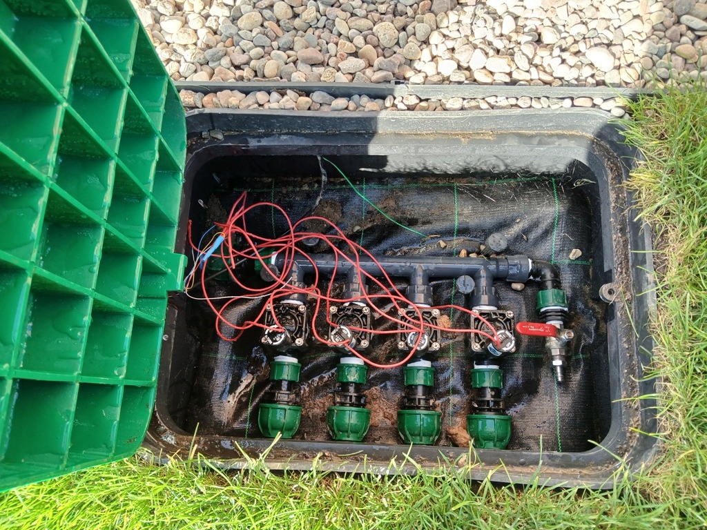 Otwarta skrzynka zaworowa z widocznymi elementami instalacji nawadniającej: elektrozawory, rury, przewody elektryczne. Na zewnątrz skrzynki trawa i kamienie ozdobne.