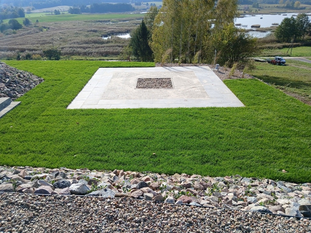 Widok z góry na świeżo założony trawnik z rolki, z centralnie umieszczonym kwadratowym paleniskiem wyłożonym kamieniami, otoczonym płytami chodnikowymi, na tle malowniczego krajobrazu z jeziorem...