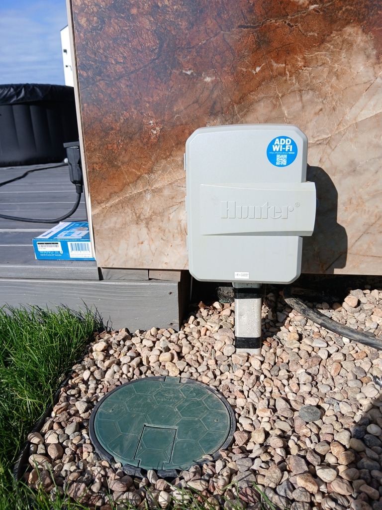 Szara skrzynka sterująca z napisem Hunter i naklejką 'Add WiFi' zamontowana na elewacji z płytek imitujących marmur, obok trawnika i pokrywy zaworu w otoczeniu drobnych kamieni.