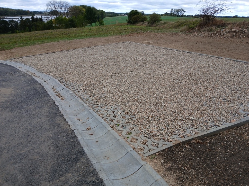 Utwardzony plac z kruszywa, ograniczony betonową obrzeżem i ażurowymi płytami, przylegający do asfaltowej drogi z betonowym korytkiem odwadniającym. W tle widoczne drzewa i pola.