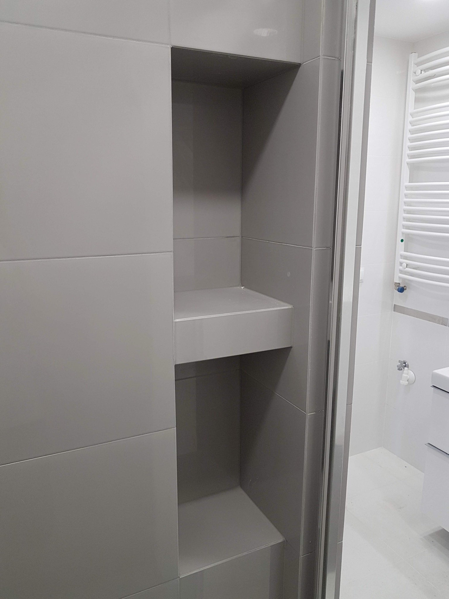 Wnęka z półkami wyłożona szarymi płytkami w nowoczesnej łazience, widoczny fragment kabiny prysznicowej i biały grzejnik drabinkowy.
