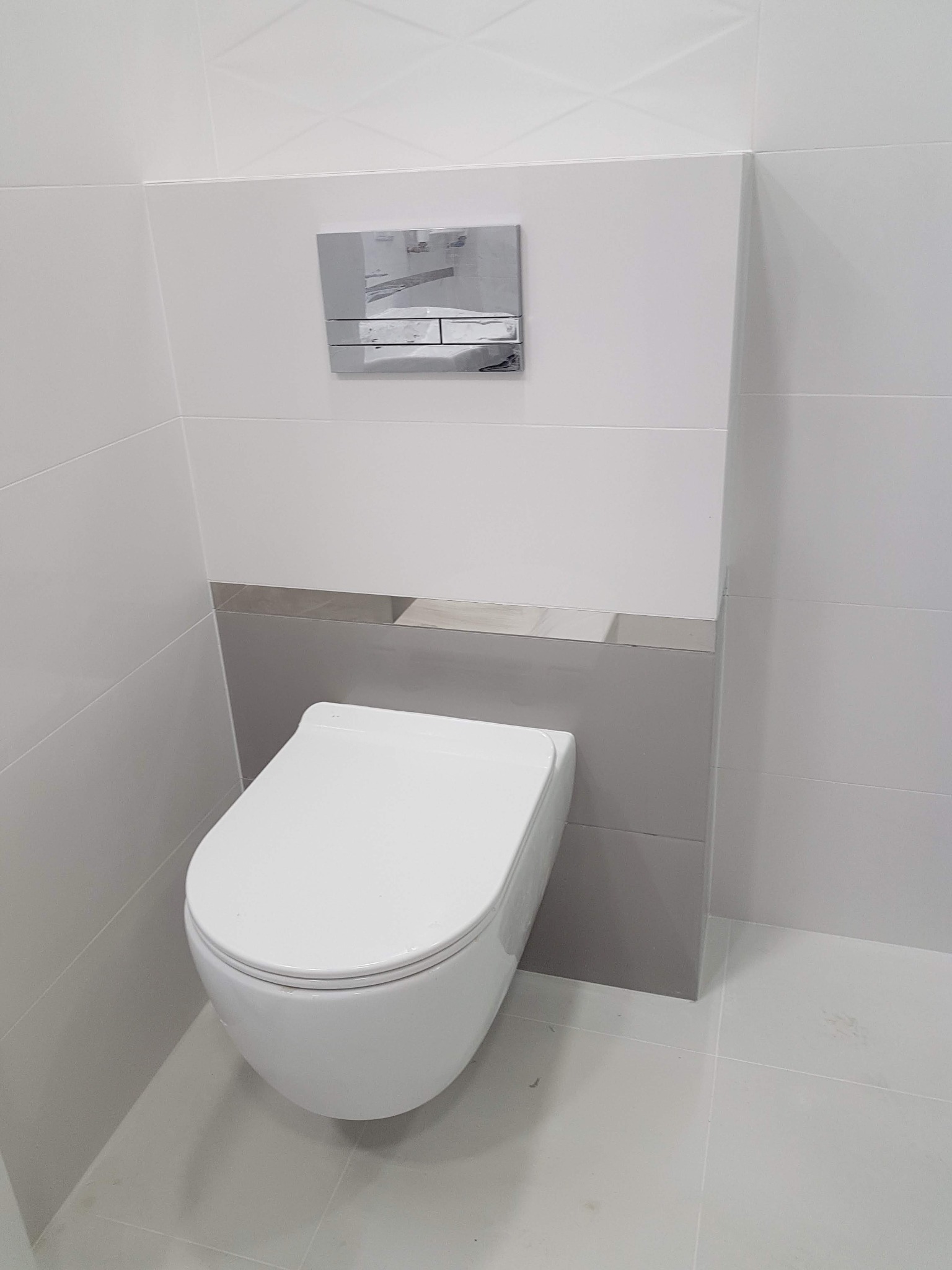 Podwieszana toaleta w nowoczesnej łazience z białymi i szarymi płytkami oraz chromowanym przyciskiem spłukującym.