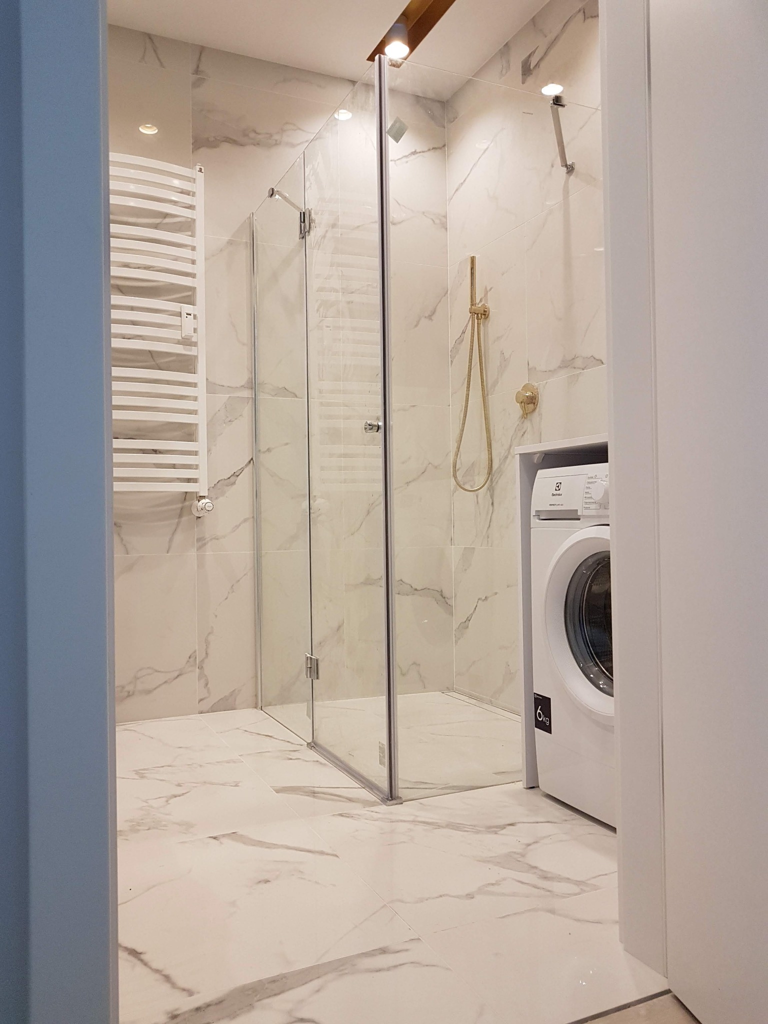 Jasna łazienka z kabiną prysznicową, grzejnikiem drabinkowym, złotą armaturą i pralką Electrolux w zabudowie, wykończona płytkami imitującymi marmur.