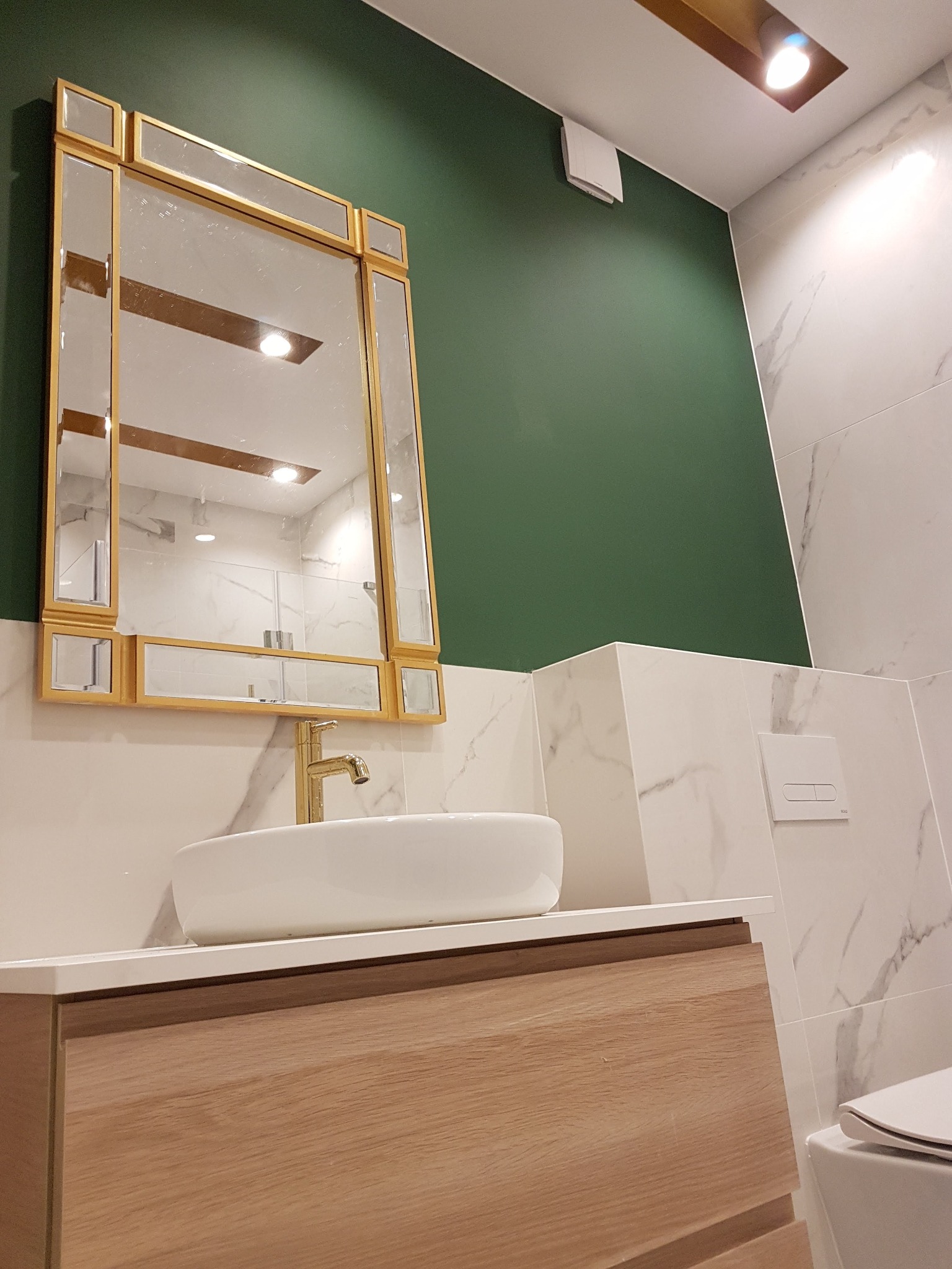 Nowoczesna łazienka z zieloną ścianą, marmurowymi płytkami i złotą armaturą, lustro w złotej ramie odbija kabinę prysznicową.