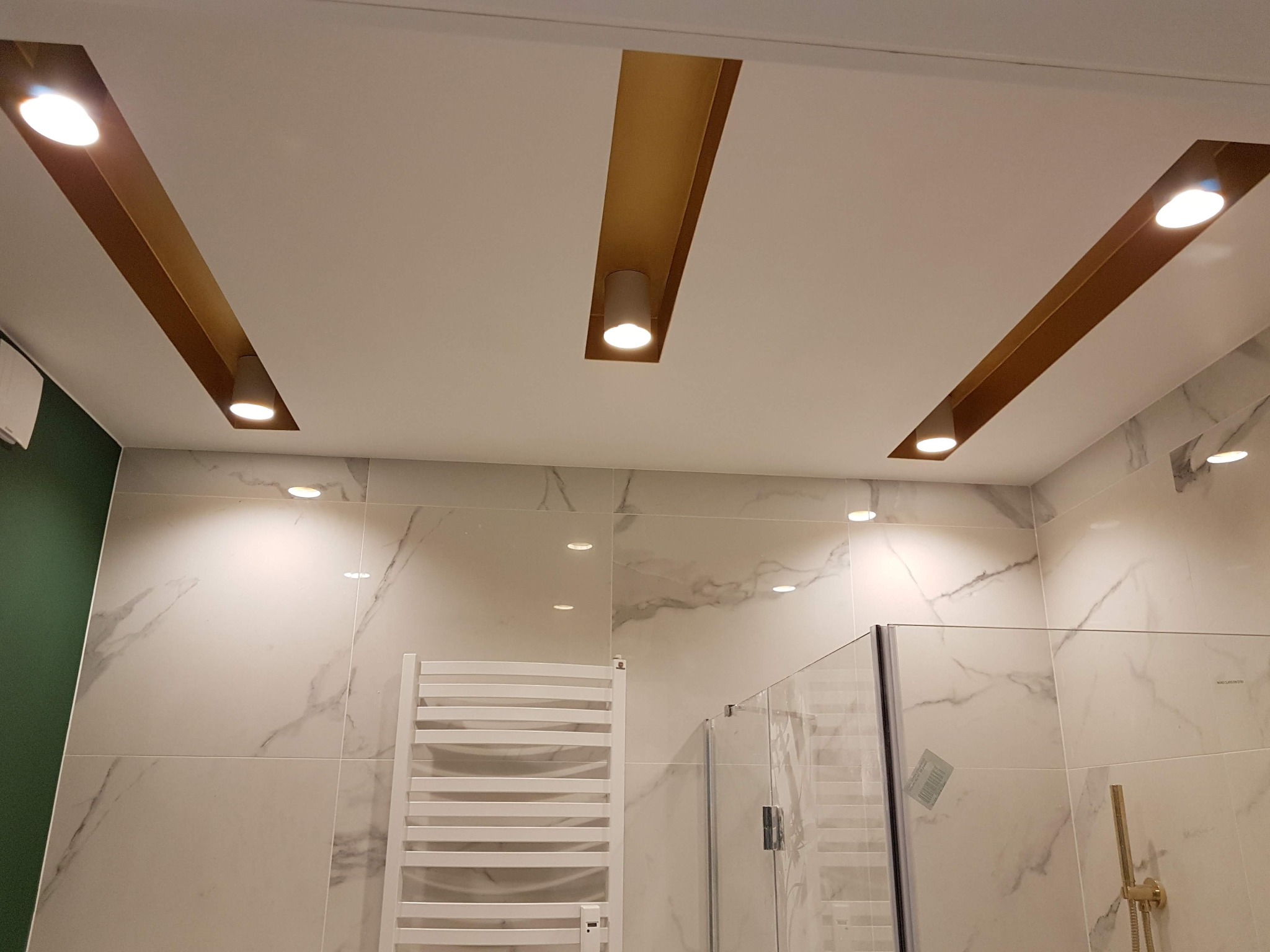 Nowoczesna łazienka z białymi płytkami imitującymi marmur, podwieszanym grzejnikiem drabinkowym i kabiną prysznicową ze złotą armaturą, oświetlona trzema podłużnymi wnękami z lampami punktowymi...