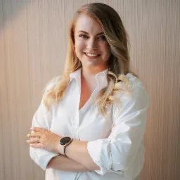 Uśmiechnięta blondynka w białej koszuli z podwiniętymi rękawami, stojąca na tle ściany o delikatnej fakturze, z założonymi rękami i zegarkiem na nadgarstku.