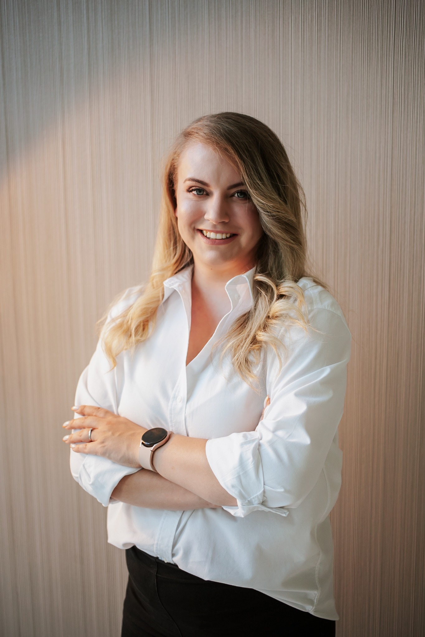 Uśmiechnięta blondynka w białej koszuli z podwiniętymi rękawami, stojąca na tle ściany o delikatnej fakturze, z założonymi rękami i zegarkiem na nadgarstku.