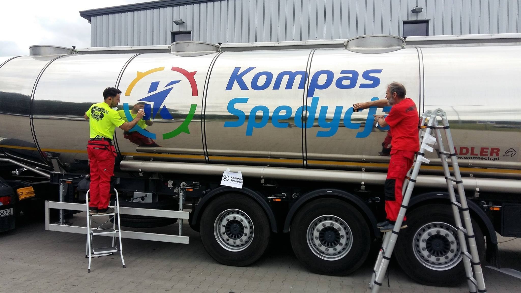Dwaj mężczyźni w czerwonych i żółtych uniformach, stoją na drabinach i czyszczą błyszczący, metalowy zbiornik ciężarówki z logo 'Kompas Spedycja'.