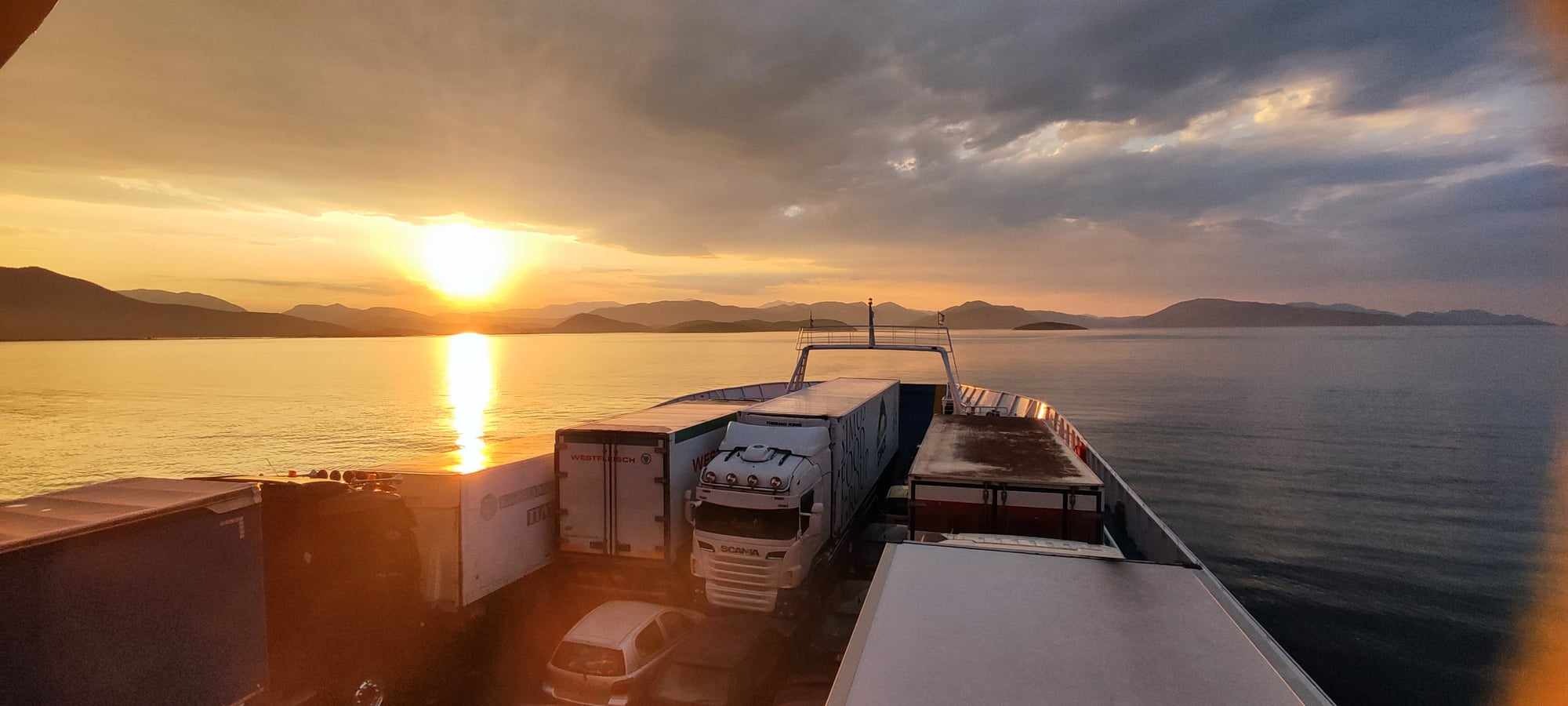 Ciężarówki i samochód osobowy na promie podczas zachodu słońca nad morzem.