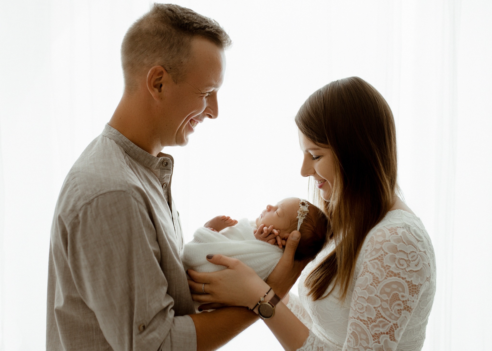 Rodzice patrzą z uśmiechem na śpiące nowonarodzone dziecko owinięte w biały kocyk, na jasnym tle okna.