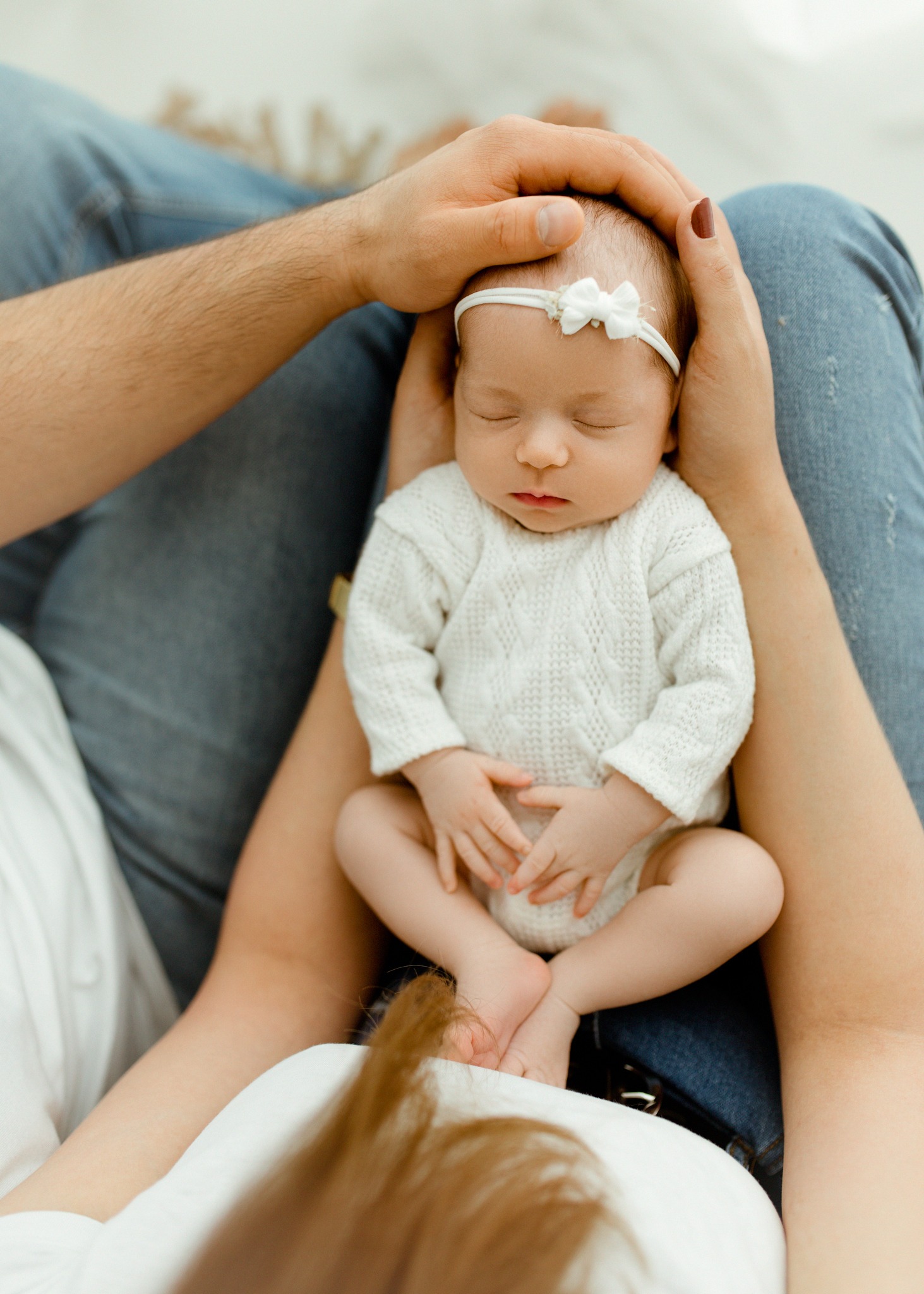 Noworodek w białym sweterku i z kokardką na głowie, trzymany w ramionach rodziców, z delikatnym oświetleniem i ciepłą kolorystyką.