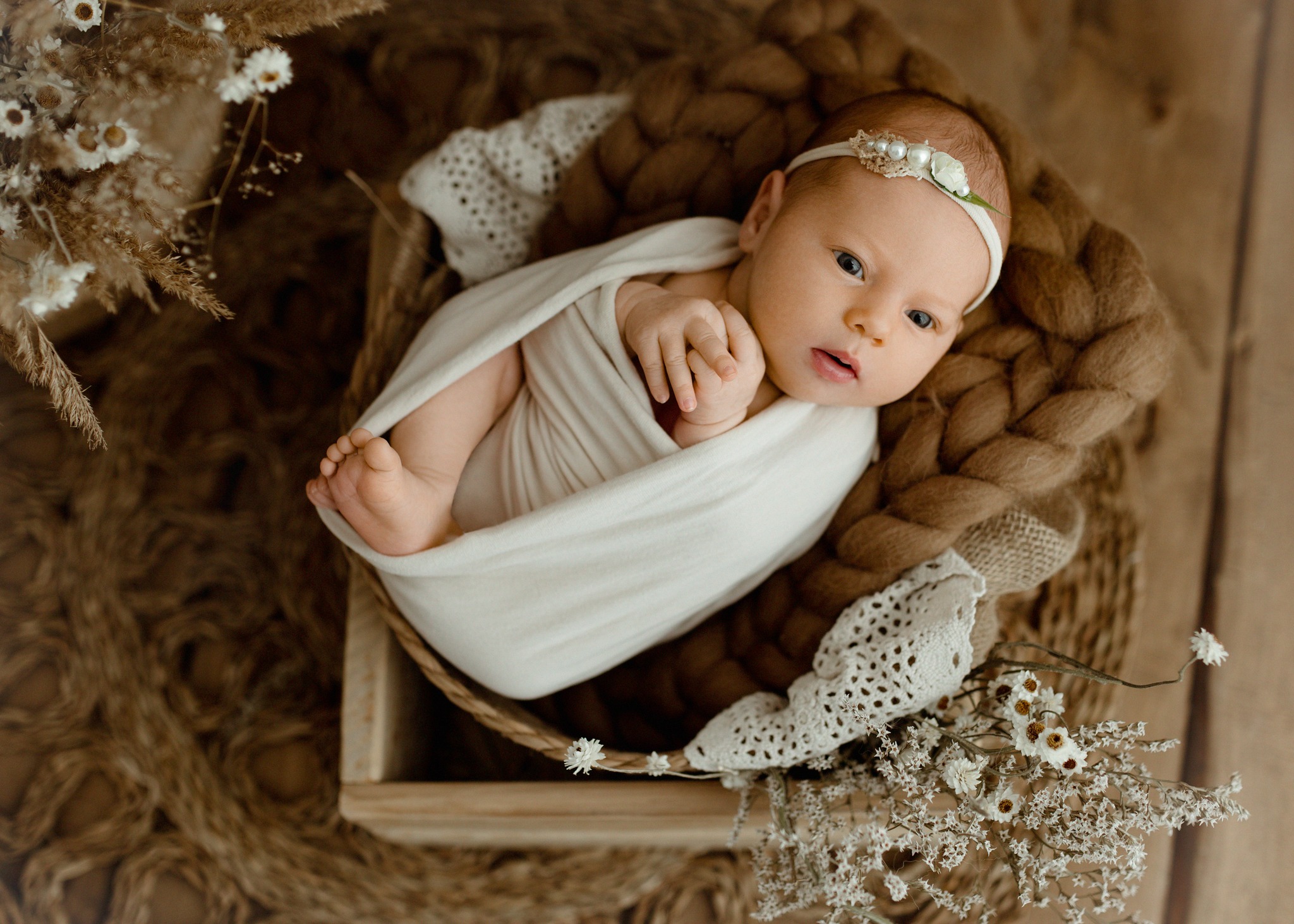 Noworodek owinięty w kremowy materiał, leżący w drewnianej skrzynce wyłożonej grubym, plecionym sznurem i koronkową serwetką, ozdobionej suszonymi kwiatami, z delikatną opaską na głowie.
