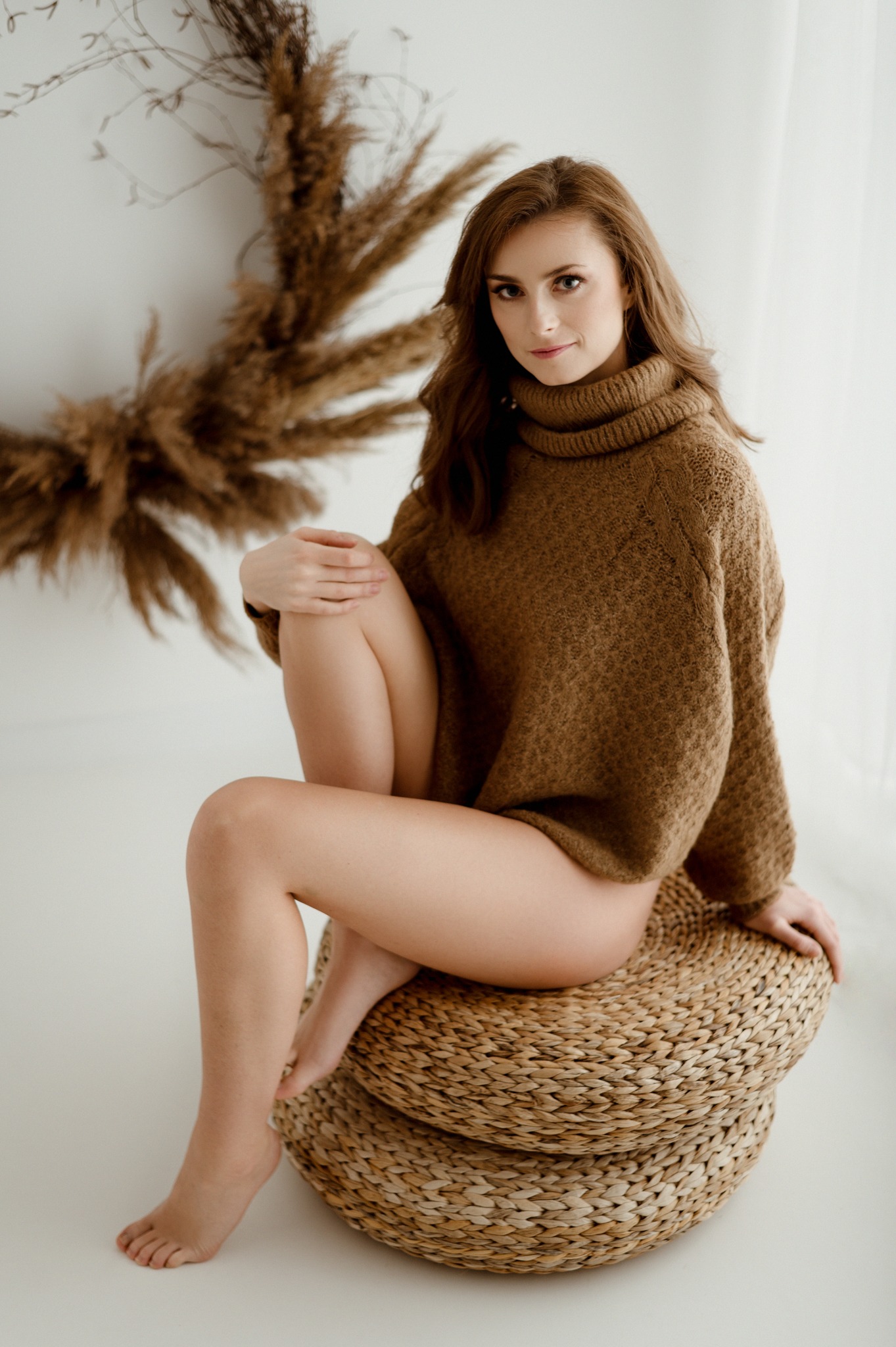 Kobieta w brązowym swetrze z golfem pozuje siedząc na okrągłym, plecionym pufie na tle minimalistycznego studia z dekoracją z suszonych traw.